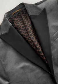 Grijze fluwelen blazer met een zwarte satijnen revers. Aan de binnenkant is er een gemêleerde voering in zwart en koper, met geometrische vormen.