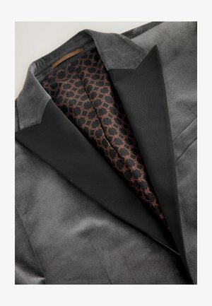 Blazer en velours gris avec un revers en satin noir. À l'intérieur, une doublure à motifs en noir et cuivre, présentant des formes géométriques.