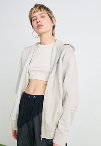 Gai bēšas krāsas zip-up sporta jaka, kas izgatavota no mīksta auduma, valkāta pāri īsai bēšas krāsas blūzei un apvienota ar melniem šortiem ar aukliņu, kuriem ir teksturēts dizains.