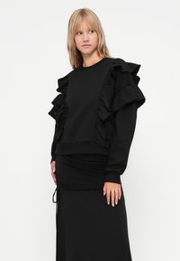 Hanorac negru cu accente volanate pe mâneci, decolteu rotund și brâu ajustat. Textură netedă, design casual, detalii în straturi.