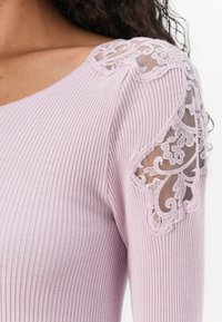 Lila geribbelde top met sheer kantaccenten op de schouders met ingewikkelde bloemmotieven. Soepele textuur, aansluitend ontwerp en lichte stretch.