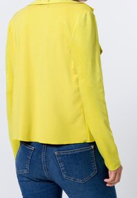 Żółta kurtka z długim rękawem, gładka w dotyku, z wykładanym kołnierzem, zestawiona z ciemnoniebieskimi dopasowanymi dżinsami z tylnymi kieszeniami.