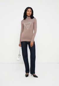 Un maglione dolcevita marrone con un logo triangolare argento al centro, abbinato a jeans denim scuri e décolleté nere a punta. Borsa bianca.
