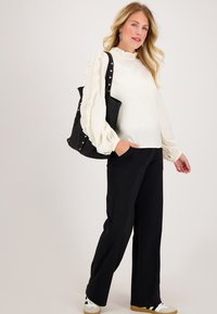 Witte, gerimpelde blouse met lange mouwen, gecombineerd met zwarte, hoge taille broek en witte sneakers. Het model houdt een zwarte schoudertas met zilveren accenten vast.