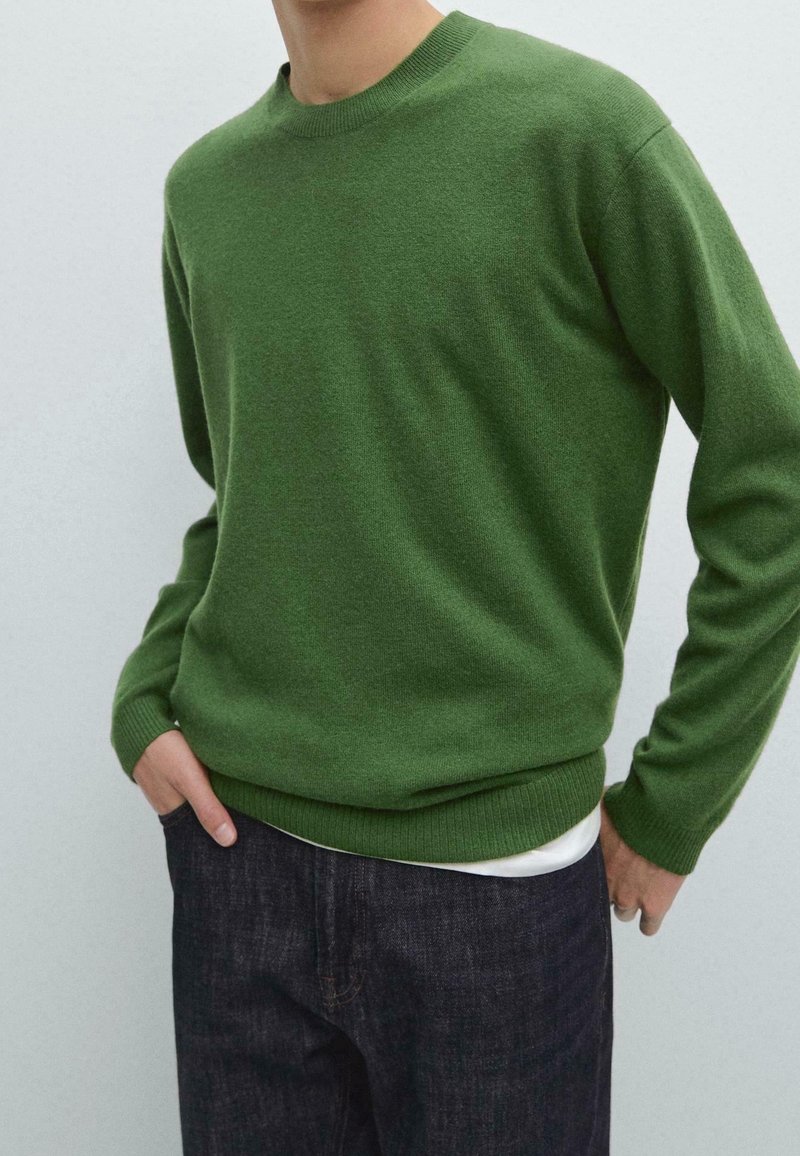 Homme portant un pull vert ajusté à col rond par-dessus une chemise blanche, associé à un jean bleu foncé, mains placées de manière décontractée dans les poches.