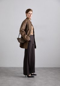 Bruine geruite oversized jas, tan top, hoge taille donkere broek en zwarte pumps met spitse neuzen. Een kleine bruine tas accentueert de outfit.