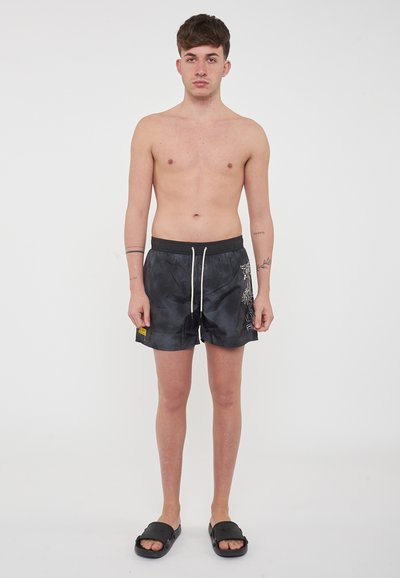 Pantaloni da nuoto neri con vita regolabile, tasche laterali e ricami floreali bianchi sulla gamba sinistra; indossati con ciabatte nere.