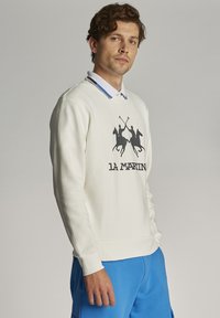 Bela majica s kapuco z grafiko dveh konjeničev in napisom "la Martin". Ima okroglo ovratnik in rebraste manšete. Nosi se preko modre srajce s kravato.