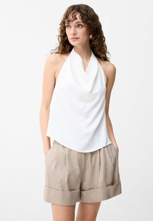 Vrouw met krullend haar, een witte haltertop en beige linnen shorts, handen in de zakken, kijkt iets opzij.
