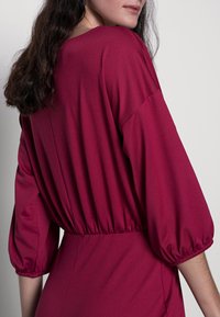 Vestido na cor de baga com um corte descontraído, apresentando mangas balão e um tecido elástico na cintura. Textura de tecido suave e design minimalista.