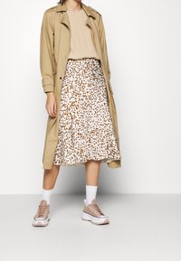 Beige trenchcoat över en ljus topp, tillsammans med en vit kjol med bruna prickar och kraftiga beiga sneakers med vita sulor.