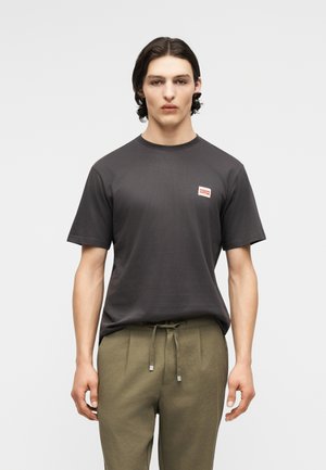 Jeune homme aux cheveux foncés portant un T-shirt gris foncé avec un petit logo rouge et blanc sur la poitrine, associé à un pantalon à cordon olive.