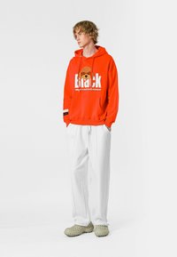 Sudadera naranja brillante con un gráfico de un oso y texto en negrita, combinada con pantalones blancos holgados y zapatos ligeros verdes tipo slip-on.