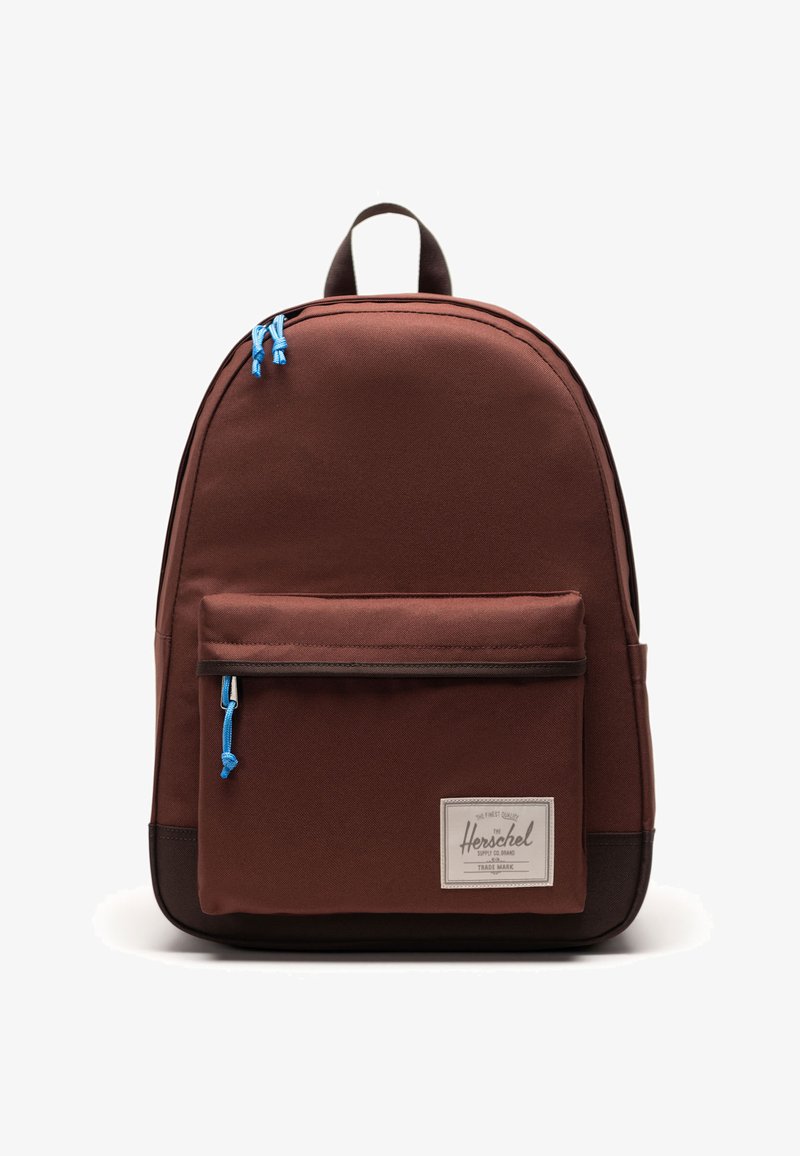 Bruine stoffen rugzak met blauwe ritsen, een voorvak en een rechthoekige logo patch. Rondvormig ontwerp en dubbele schouderbanden.
