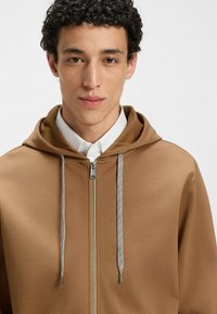 Brown zip-up hoodie featuring a smooth texture, contrasting silver zipper, and white drawstrings with metallic tips.