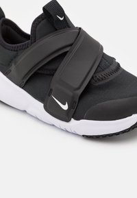 Black athletic shoe featuring a fabric upper, a thick black strap, white sole, and signature logo. Textured accents on the upper.