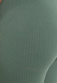 Zelené legíny s vroubkovaným vzorem vyrobené z elastického materiálu, s hladkým povrchem a jemným detailem švu. Záběr zblízka zdůrazňuje materiál.