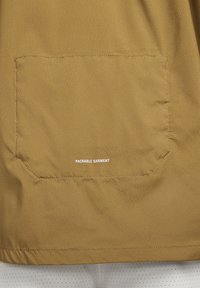 Gros plan d’une poche en tissu kaki avec de petites perforations et un texte blanc indiquant « PACKABLE GARMENT » centré sur la poche.