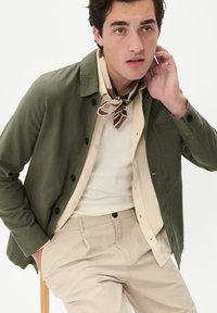 Olivgrüne, knöpfbare Jacke mit strukturierter Oberfläche, getragen über einem beigefarbenen, gerippten Shirt, kombiniert mit hellbeigen Hosen und einem braunen, gemusterten Schal.