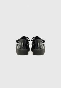 Chaussures en cuir verni noir avec une semelle en caoutchouc texturé, ornées de nœuds à l'arrière et d'une forme arrondie lisse.