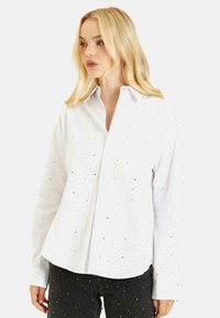 Camicia bianca a maniche lunghe con colletto a punta, decorata con paillettes argento e vestibilità rilassata. Presenta polsini con bottoni e orlo curvo.