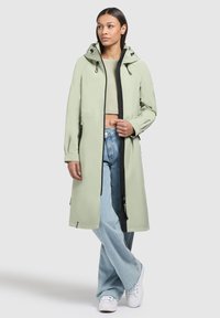 Light green waterproof jacket with a hood, black zipper, and drawstrings. Tailored fit paired with loose blue denim jeans and white sneakers.