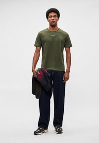 Λαδί βαμβακερό μπλουζάκι με κοντά μανίκια, φορεμένο με φαρδιά σκούρα μπλε τζιν. Το μοντέλο κρατάει ένα σκουρόχρωμο πουλόβερ με σχέδια και φοράει πολύχρωμα αθλητικά παπούτσια.