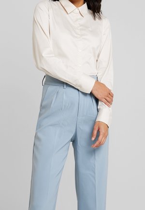 Camicia button-up color crema chiaro con colletto, abbinata a pantaloni a vita alta blu chiaro con pieghe e una texture liscia.