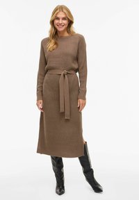 Brązowa dzianinowa sukienka z okrągłym dekoltem, długimi rękawami, paskiem wiązanym w talii oraz rozcięciami po bokach, zestawiona z czarnymi kozakami za kolano.