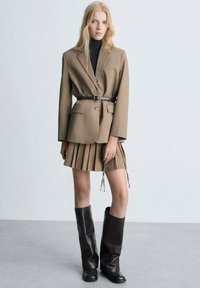 Rjav krojen blazer z pasom, v kombinaciji s plisirano krilo v houndstooth vzoru in rjavimi usnjenimi škornji do kolen. Gladka tekstura, minimalno okraševanje.
