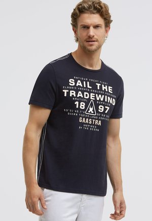 Mann trägt ein marineblaues T-Shirt mit weißem nautischem Text und Symbolen, kombiniert mit weißen Hosen, steht vor einem schlichten hellen Hintergrund.