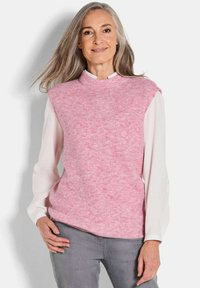 Lächelnde Frau mit langen grauen Haaren, die einen rosa ärmellosen Strickpullover über einer weißen Bluse und grauen Hosen trägt, steht vor einer weißen Wand.