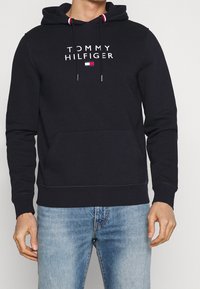 Marineblauer Hoodie aus Baumwollmischung, mit einer Fronttasche, Kordelzugkapuze und dem weißen "TOMMY HILFIGER"-Logo mit kleinem Logoakzent.