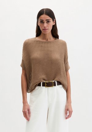 Woman wearing a loose, short-sleeve, knitted beige top tucked into high-waisted white pants with a dark brown belt.