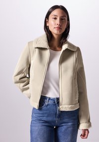 Beige läderjacka med en plysch, krämfärgad fårskinnskrage och manschetter; har en framdragbar dragkedja och en kort passform. Blå jeans bärs.
