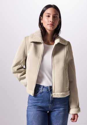 Veste en cuir beige avec un col et des poignets en shearling crème duveteux ; elle présente une fermeture éclair à l'avant et une coupe courte. Un jean bleu est porté.