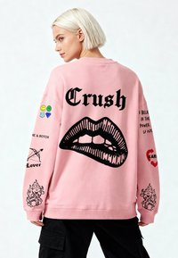 Felpa oversize rosa con testo grafico nero "Crush" e design di labbra sul retro, caratterizzata da schemi unici e applicazioni colorate sulle maniche.