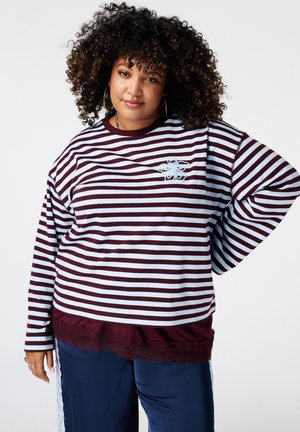 Femme aux cheveux bouclés portant un haut à manches longues rayé bordeaux et blanc avec un détail floral, un pantalon bleu marine et un bord en dentelle, debout avec la main sur la hanche.