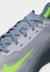 Κοντινή λήψη ανοιχτόγριου παπουτσιού Nike για τρέξιμο με γκρι κορδόνια και λογότυπο Swoosh σε νέον πράσινο χρώμα πάνω σε υφασμένο ύφασμα.