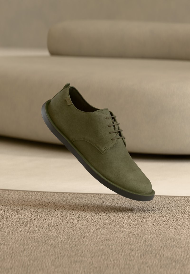 Πράσινο ελιάς σουέτ παπούτσι με κορδόνια, στρογγυλερή μύτη και μαύρη ελαστική σόλα, να αιωρείται πάνω από μια υφασμένη μπεζ επιφάνεια.