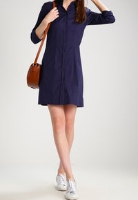 Marinblå skjortklänning i lätt tyg. Har krage, långa ärmar och slutar ovanför knäet. Kommer ihop med vita sneakers.