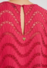 Dos d'une robe en dentelle rose avec des motifs floraux, une petite ouverture en forme de trou de serrure et une étiquette dorée en forme de cœur portant l'inscription Twinset Milano.