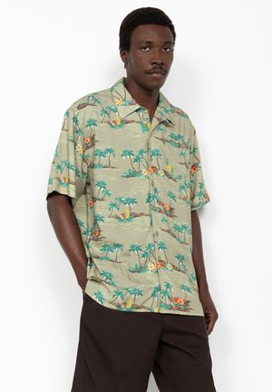 Homme portant une chemise à manches courtes boutonnée avec un imprimé de palmiers tropicaux verts et de voiliers, assortie à un pantalon foncé.