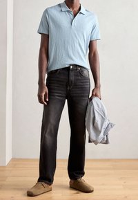 Ljusblå pikétröja med korta ärmar, mörka denimjeans och beige träskor. Randig skjorta hålls i vänster hand. Enkel design, mjuka texturer.