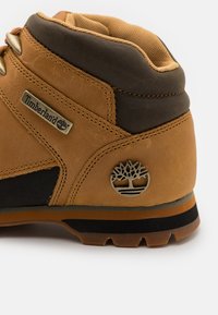 Brune skinnstøvel med en tan og mørk brun finish, med metalløyler og en sølvtrær-logo på siden. Teksturert detaljering gjennom hele støvelen.