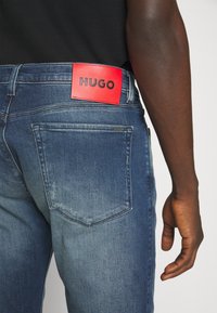 Vue de dos d'une personne portant un jean bleu avec une étiquette rouge portant la mention "HUGO" au-dessus de la poche arrière droite et une chemise noire.