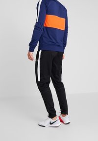 Sweatshirt de sport marine avec une bande horizontale orange et des détails blancs, associé à un bas de survêtement noir avec des rayures blanches sur les côtés et des chaussures de sport.