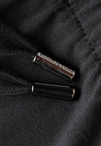 Schwarzer Stoff mit strukturiertem Gewebe, der an beiden Enden Zugbänder hat. An einem Ende befindet sich eine silberne Metallspitze, die mit "intimissimi" graviert ist.