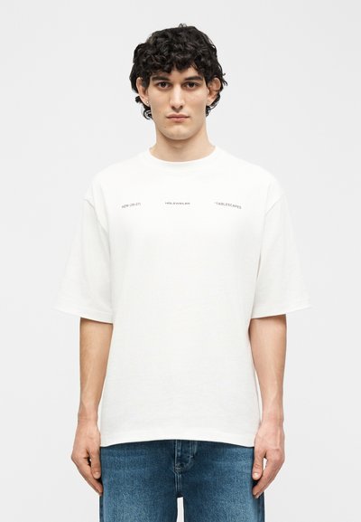 Νεαρός με σγουρά μαλλιά που φορά λευκό φαρδύ μπλουζάκι με μικρό μαύρο κείμενο και μπλε τζιν, στέκεται μπροστά σε απλό φωτεινό φόντο.