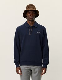 Jūras zila garām piedurknēm polo krekls ar apkakli, ar logo uz krūtīm un ribotām piedurknēm. Kombinēts ar brūnu kausa formā cepuri.